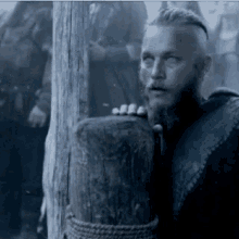 a man with a beard is leaning against a wooden post with his hand on it .
