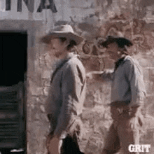 two men are standing next to each other in front of a stone wall .