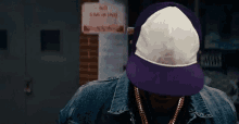a man wearing a purple hat and a denim jacket stands in front of a no smoking sign .
