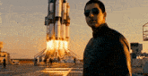 a man wearing sunglasses stands in front of a rocket that is being launched