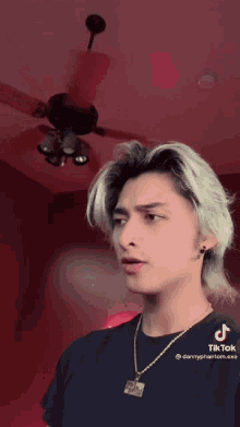 a man wearing a black shirt and a gold necklace is standing in front of a ceiling fan in a room .