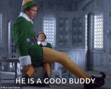 a man in a green elf costume sits on a chair with the words he is a good buddy written below him