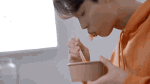 a young man in an orange hoodie is eating noodles with chopsticks from a bowl .