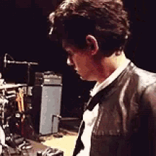 a man in a black leather jacket is standing in front of a drum set .