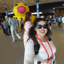 a woman wearing sunglasses stands in front of a large yellow duck with a pink beak
