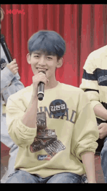 a young man with blue hair is holding a microphone and wearing a yellow sweatshirt that says now
