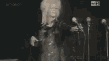 a woman with blonde hair and a leather jacket is standing in front of a black wall .
