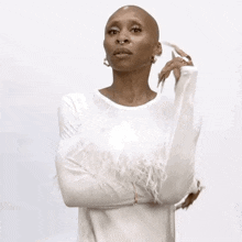 a bald woman wearing a white feathered sweater is standing with her arms crossed and looking at the camera .