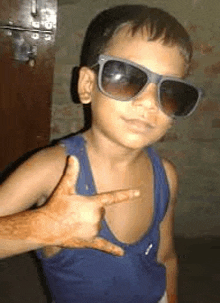 a young boy wearing sunglasses and a blue tank top is pointing to his chest .