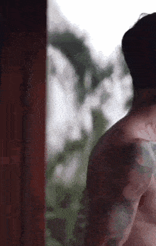 a shirtless man with tattoos on his back is standing in front of a window looking out .