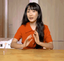 a woman in an orange shirt sits at a table with her hands together