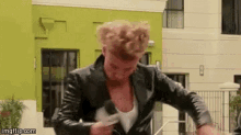 a man in a leather jacket is standing in front of a green building holding a microphone .