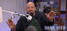 a bald man is sitting in front of a microphone with his arms outstretched in front of a dark web logo