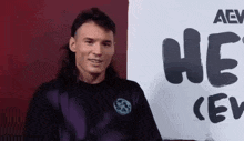 a man with long hair and a mullet is standing in front of a sign that says `` he 's crew '' .