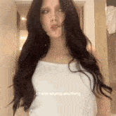 a woman with long dark hair is wearing a white tank top and making a face .