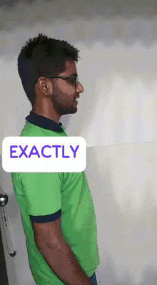 a man wearing glasses and a green shirt is standing in front of a white wall and a sign that says exactly