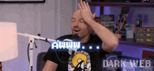 a man is sitting in front of a microphone with his hand on his head and the words awww on the screen behind him .