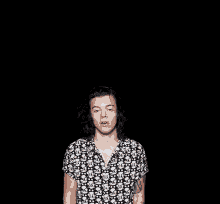 harry styles is wearing a black and white shirt with skulls on it and is standing in the dark with his eyes closed .