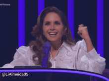 a woman in a white shirt stands in front of a blue microphone with the hashtag lialmeida05