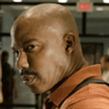 a bald man with a mustache is wearing an orange shirt and looking at the camera .