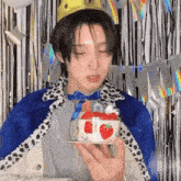 a young man wearing a crown and a cape is holding a small cake with strawberries on it .