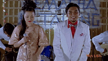 a man in a white suit stands next to a woman with a microphone in front of a man laksa sign