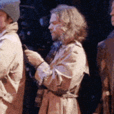 a man in a trench coat is holding a cell phone while standing next to another man .