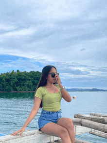 a woman wearing sunglasses sits on a wooden railing overlooking the ocean