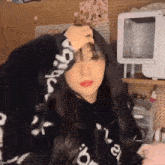 a girl in a black hoodie is standing in a room holding her head .