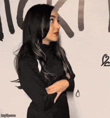 a woman in a black dress is standing in front of a wall with her arms crossed and making a funny face .