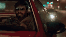 a man with a beard and sunglasses is driving a red car .