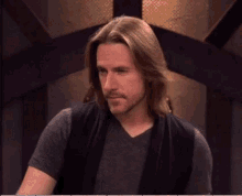 a man with long hair and a beard is wearing a gray shirt and black vest