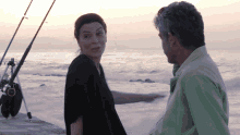 a man and a woman are standing in front of a body of water with fishing rods in the background