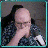 a bald man wearing glasses and a blue shirt is covering his mouth with his hand .