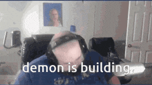 a bald man wearing headphones is sitting in front of a computer and says demon is building .