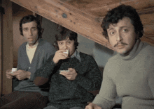 a man with a mustache is drinking from a cup while two other men look on