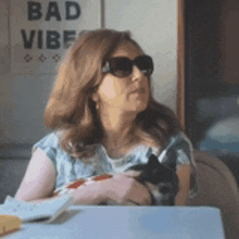 a woman wearing sunglasses is sitting at a table holding a small black cat .