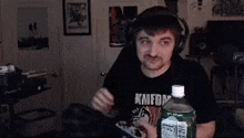 a man wearing headphones and a kmfdm shirt is sitting at a table with a bottle of soda .