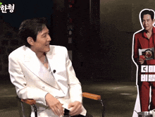 a man in a white jacket sits in front of a cardboard cut out of a man in a red suit