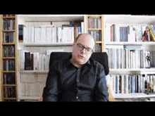 a bald man wearing glasses is sitting in front of a bookshelf filled with books .