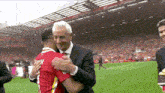 a man in a suit and tie is hugging a man in a red shirt on a soccer field