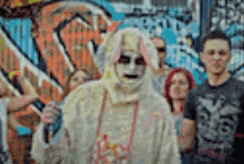 a group of people are standing in front of a graffiti wall and one of them is wearing a zombie costume .