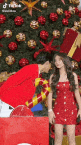 a man and a woman are standing in front of a christmas tree . the woman is wearing a red dress .