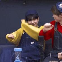 a man wearing overalls and a yellow sweater is sitting next to another man