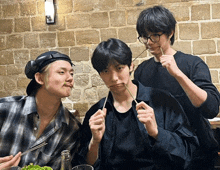 three young men are eating food with chopsticks in front of a stone wall
