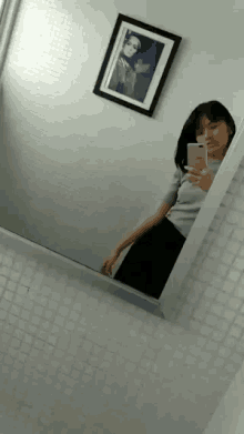 a woman is taking a selfie in front of a mirror with a picture on the wall behind her .
