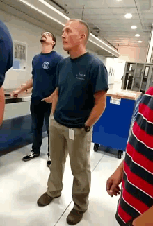 a man in a blue shirt with the letter o on it stands in a room with other people