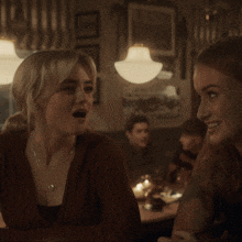 two women are sitting at a table with the words tell me everything behind them