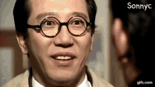 a man wearing glasses and a tan jacket is smiling and looking at another man .