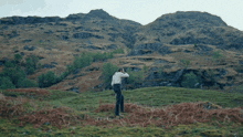 a man in a white shirt and suspenders is standing in a field with mountains in the background